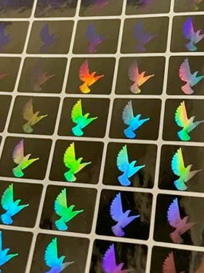 A sheet of many small holographic stickers that portray an dove in flight reflecting rainbow colors on a silvery background.