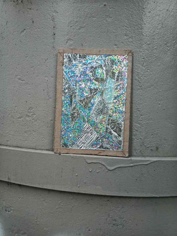 A close-up view of a small silvery glittery painting resting on a metal bar that wraps around a concrete column that is painted light grey.